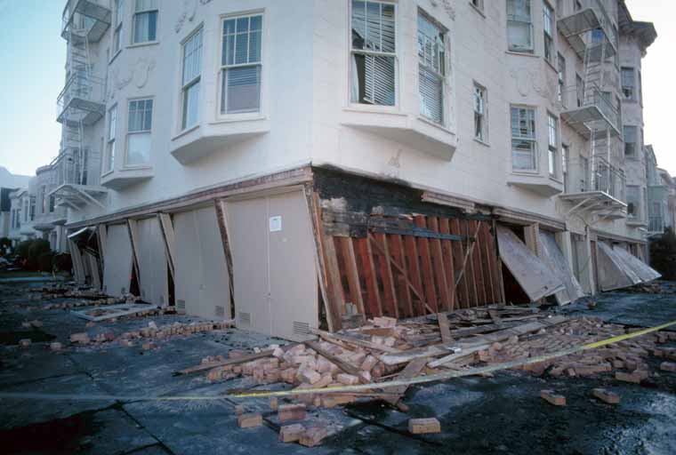 A collapsed soft-story apartment building in San Francisco's Marina district after the 1989 Loma Prieta earthquake: the open parking ground floor has given way, leaving the largely intact upper floors sunken and tilted, with exposed wooden studs, brick and concrete debris on the sidewalk, and yellow caution tape across the street.
